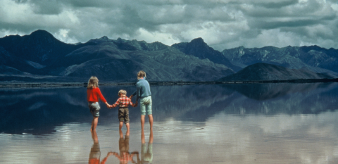 Three children standing in a shallow glassy lake before a mountain range.