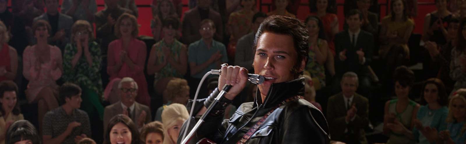 Austin Butler as Elvis Presley, dressed in a black leather suit and singing in front of a TV audience in the film ELVIS