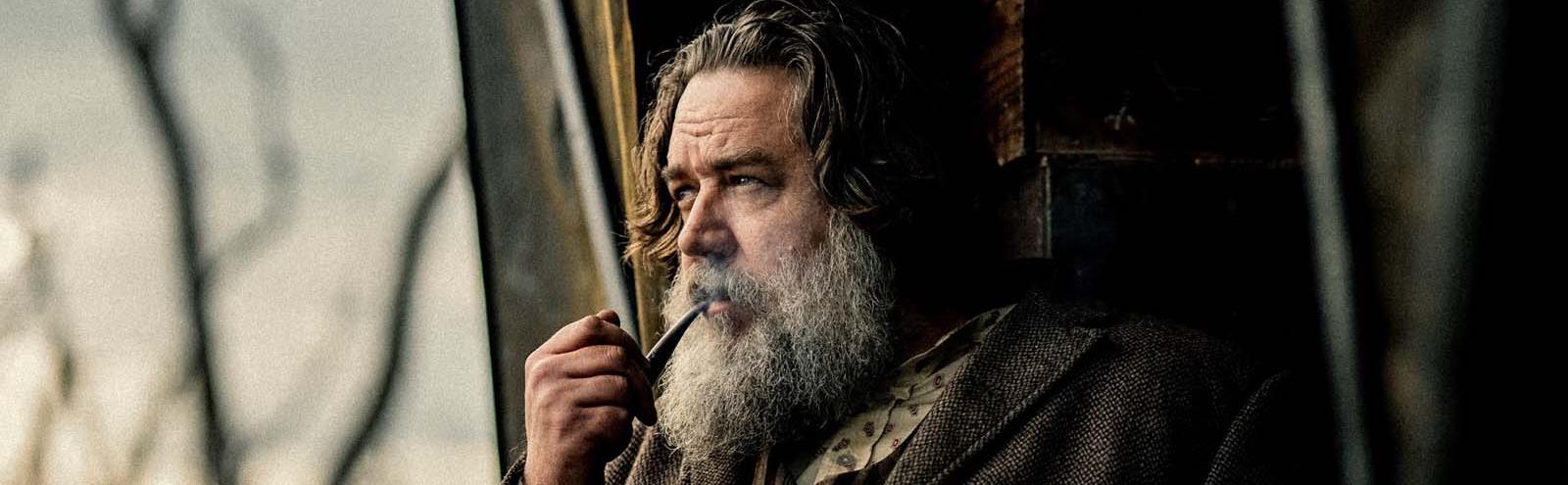 Russell Crowe in 19th century clothes looking pensively out a window while smoking a pipe in the film True History of the Kelly Gang