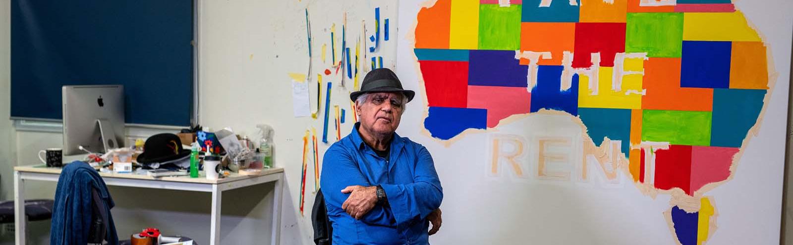 Artist Richard Bell seated in his studio in front of one of his works: a coloured Indigenous map of Australia with the words PAYE THE RENT across it