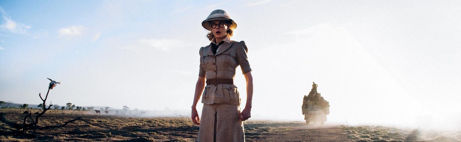 Wide shot of actor Nicole Kidman in a safari-style outfit consisting of a belted shirt and skirt and hat standing in the outback.