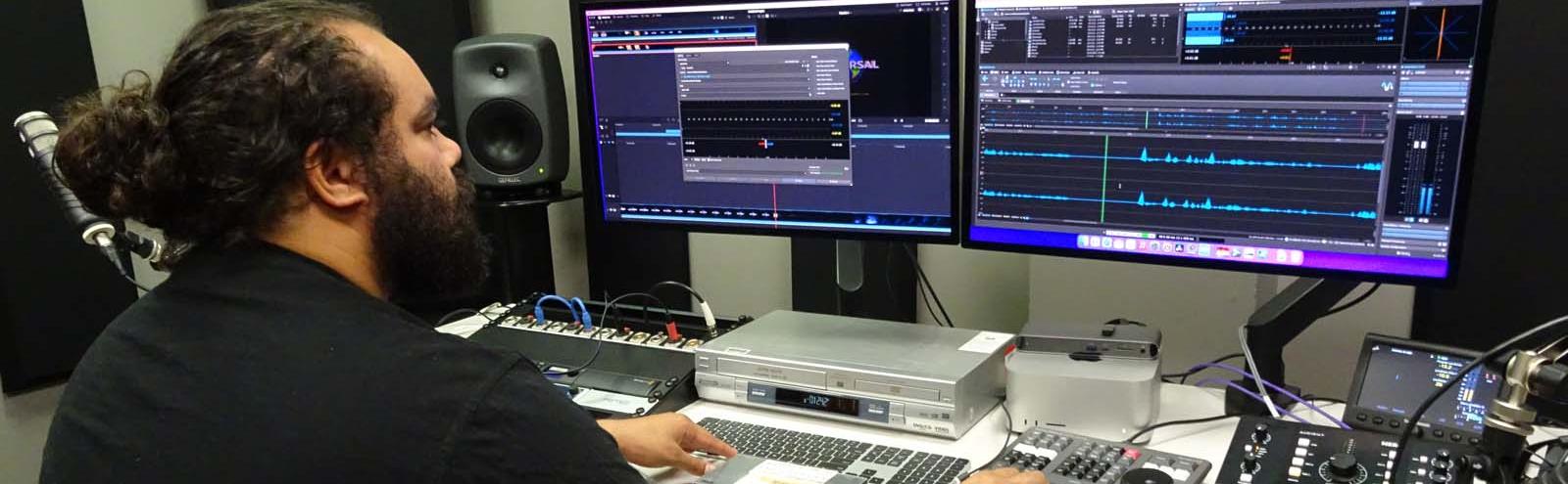 An Aboriginal man seated at a computer in front of monitors and other equipment in a recording studio