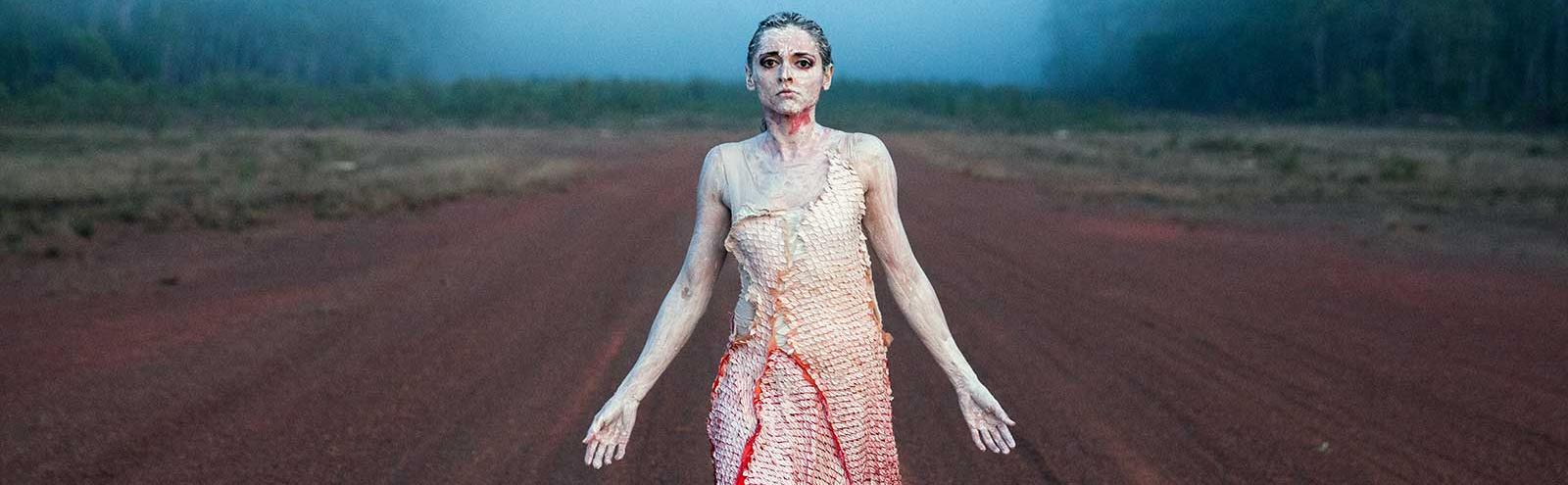 A woman wearing pale face and body paint and a pale dress with red shadings stands on a dusty dirt road as smoke rises on the horizon.