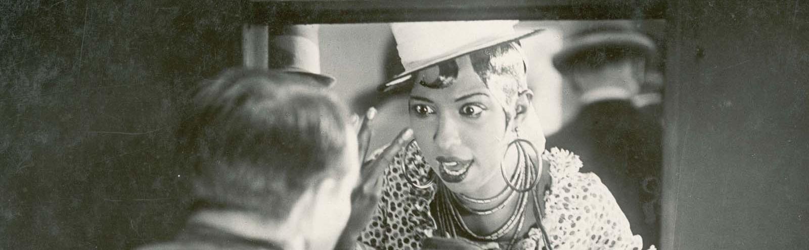 Josephine Baker holds up two fingers in an animated discussion with a man through a ticket window.