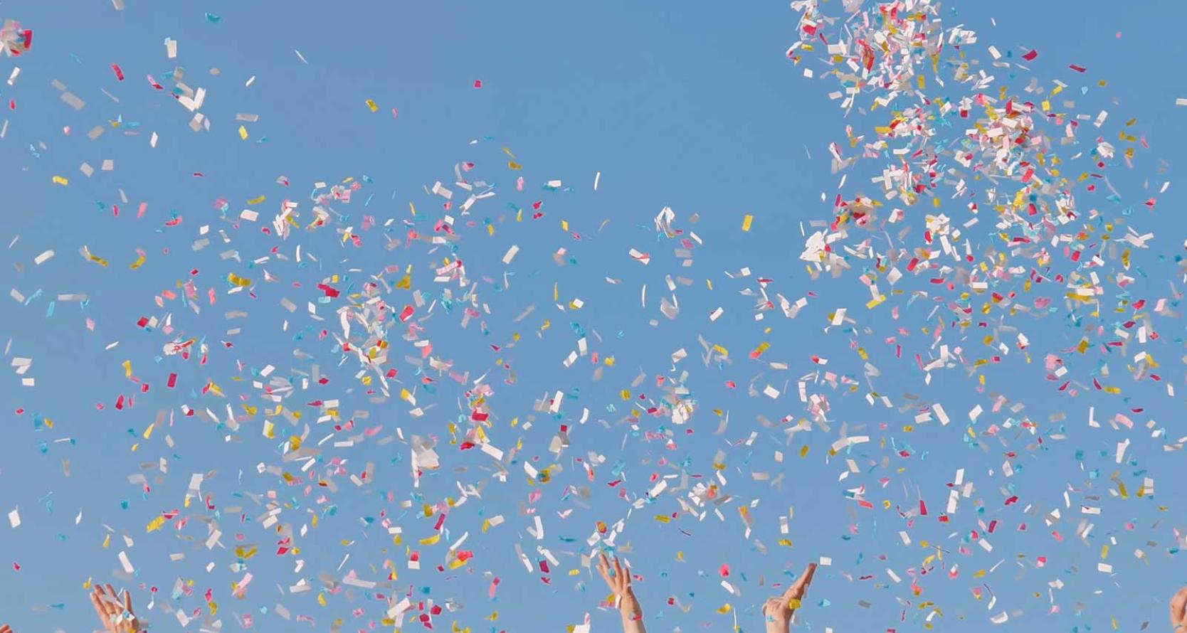 Confetti against a blue sky with hands reaching up towards it. 