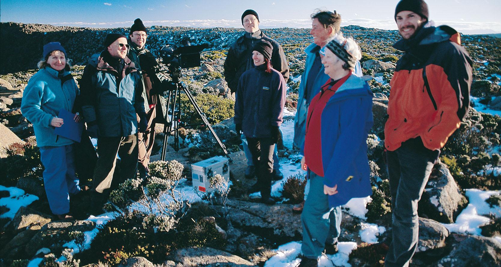 Eight adults in warm hiking clothes stand in a group on a rocky plateau. There are patches of snow on the ground and one of the men stands behind a camera tripod.