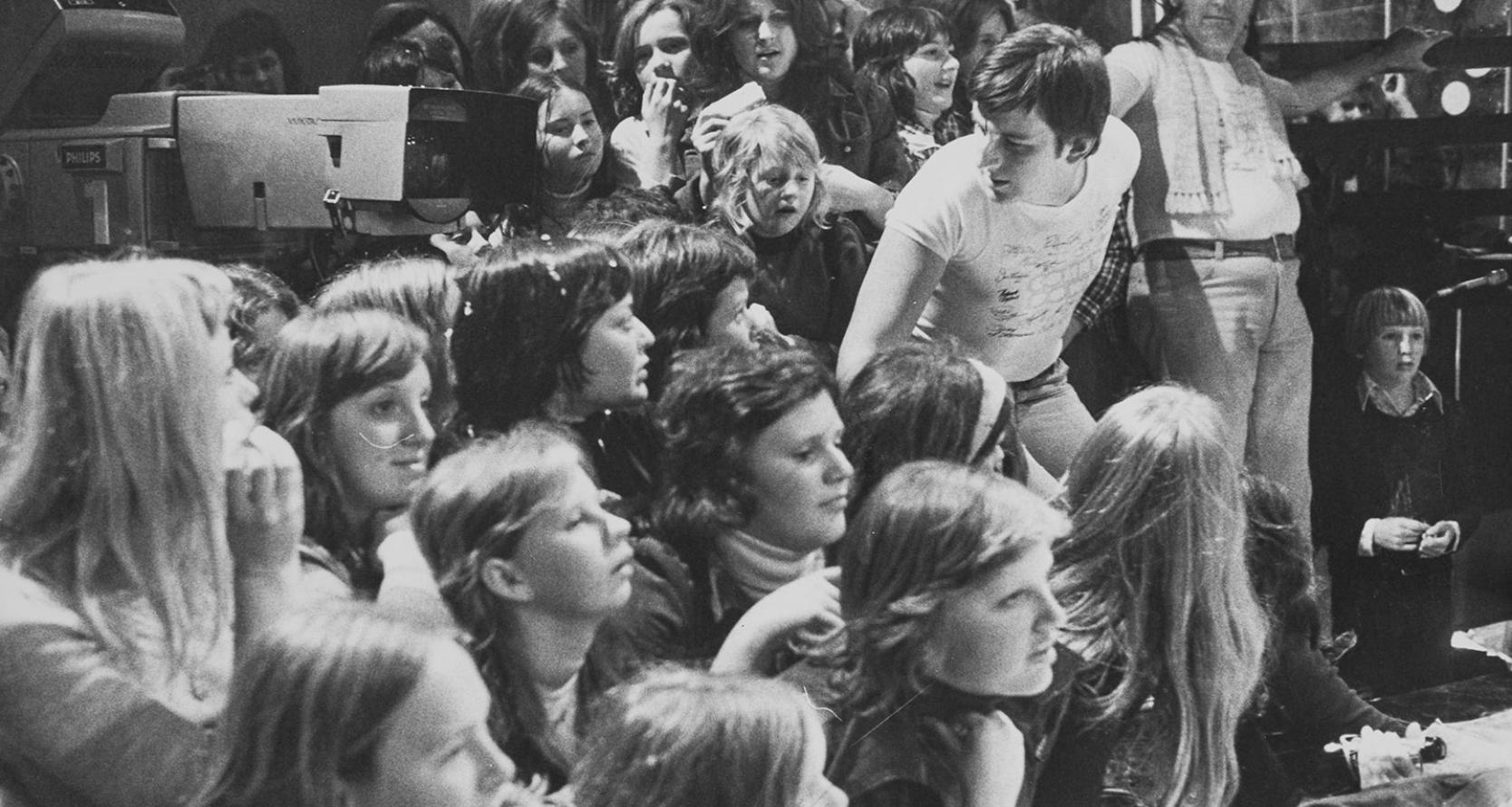 A crowd of teenagers in a TV studio sitting behind the cameraman.