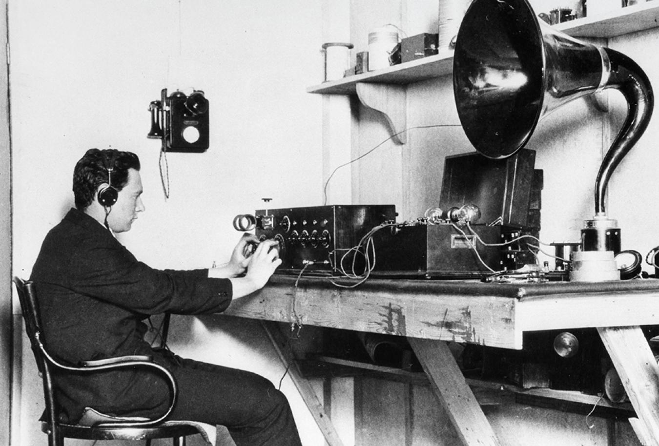 A man sitting on a desk with headphones on. There is radio and other audio equipment on the desk. c1920s