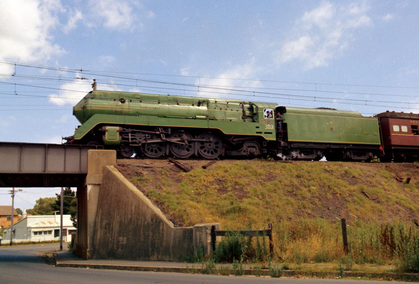 A 3801 steam locomotive pulling a passenger train on a raised railway track above a grass embankment. The train is about to cross a railway bridge