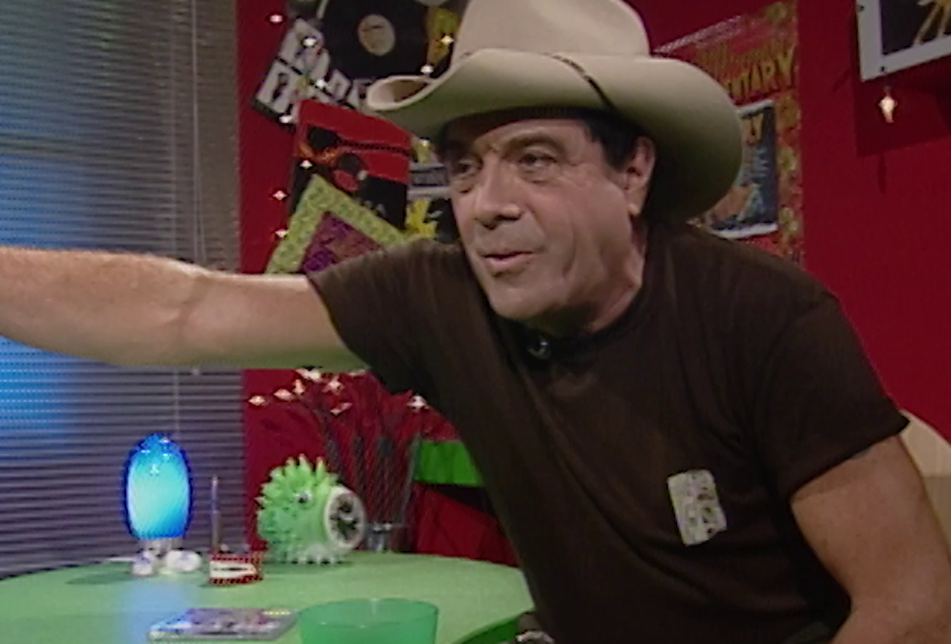 Molly Meldrum hosting music television show Countdown in the 1980s. He is seated at a table with his arm out, wearing an akubra hat and a black t-shirt