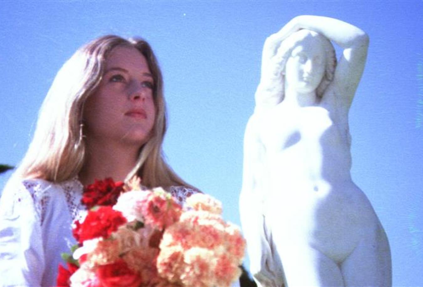 Miranda holds a bunch of colourful flowers and stands next to a statue of a woman in an outtake from Picnic at Hanging Rock.