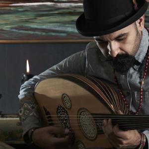 Joseph Tawadros wearing a black bowler hat and seated on a divan, playing an oud