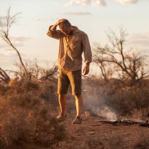 Guy Pearce and Robert Pattinson by a smouldering bushfire in an outback setting either at dusk or early dawn. Pattinson is crouching while Pearce stands apart, looking down, with hand on his head