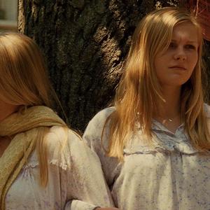 Four young women with long blonde hair wearing pale nighties and standing outside around a tree during the daytime, with mournful expressions