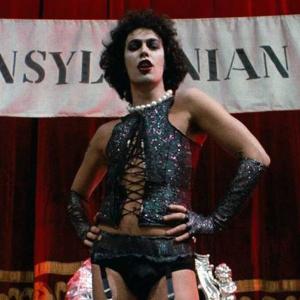 A transgender woman wears a glittery corset and white pearl necklace and stands on stage in front a banner reading Transylvanian Convention