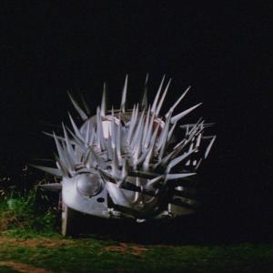 A silver Volkswagen covered in spikes like an echidna, and parked on a country road at night