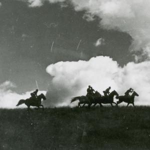 Five horses with riders seen in silhouette riding across a flat landscape beneath a cloudy sky