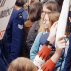 Two women standing in a crowd of protesters and police. A placard near them reads "Drop all charges against the 53".