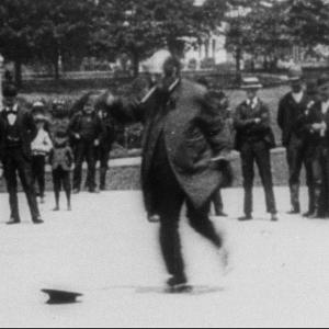 A man skating in a park in front of a crowd of people, circa 1896.