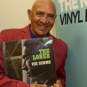 Vic Simms holding up a copy of his vinyl record "The Loner" and standing in front a banner advertising the NFSA's Vinyl Lounge