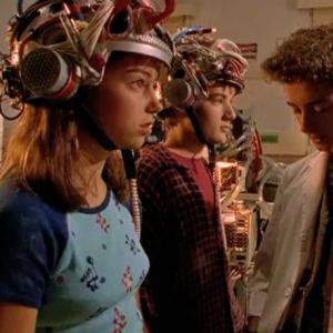 Two young teenagers stand wearing mind-control helmets and staring blankly while another teenager stands talking to them.