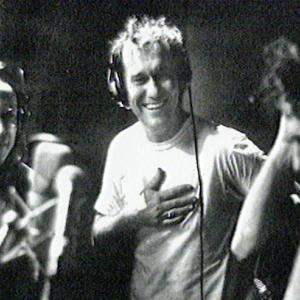 Phil Small, Jimmy Barnes and Ian Moss from the band Cold Chisel standing in a recording studio in front of microphones and all wearing headphones. 