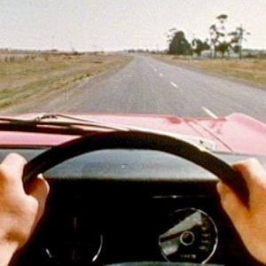 Point of view of a man driving a vintage XT Falcon on a straight stretch of road. We see his hands on the steering wheel.