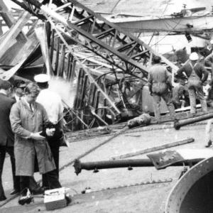 A black and white photo of the scene of the West Gate Bridge disaster. It shows investigators and bridge workers alongside the metal wreckage of the bridge.