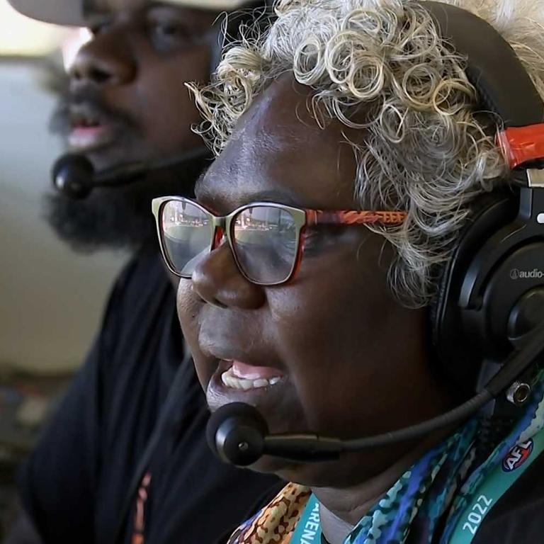 A close up of a First Nations woman with curly grey hair and glasses with headphones and a microphone on her head. There is a younger First Nations man in a hat sitting beside her in the background.