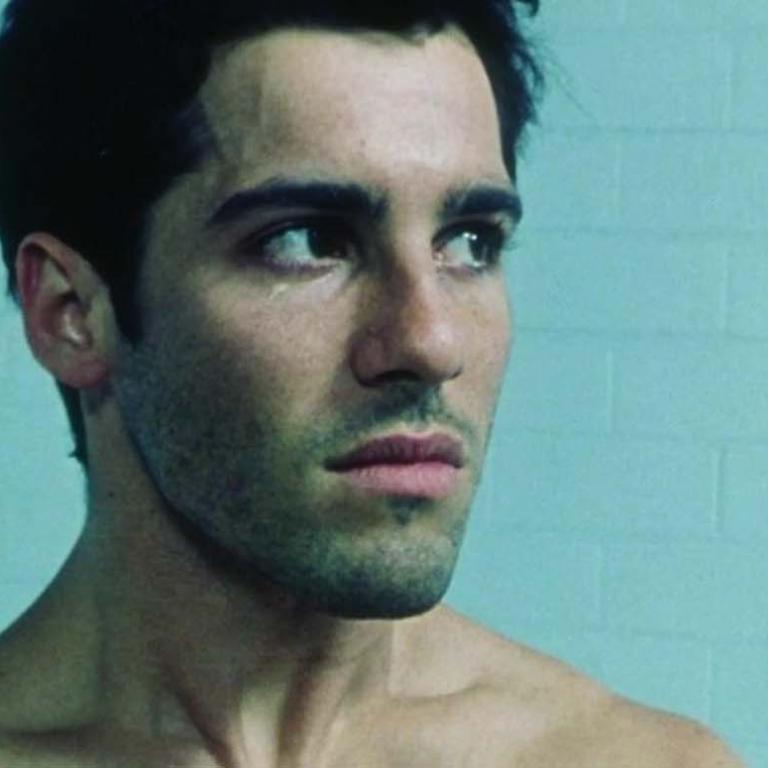 A shirtless Greek-Australian man stands in front of a tiled wall