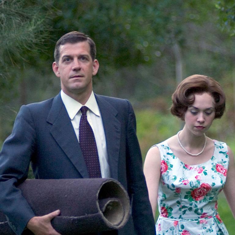 A man, dressed in a suit and tie, walks hand in hand with a woman, dressed in a floral frock, through a grassy, wooded area.