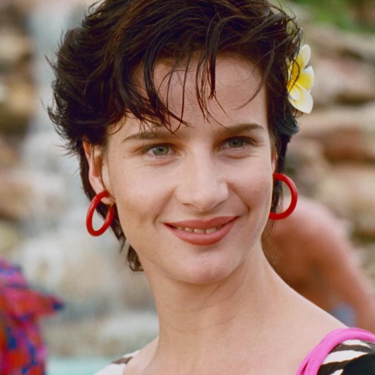 Head and shoulders shot of Rachel Griffiths in a scene from Muriel's Wedding. She's wearing bright red earrings and lipstick and has a frangipani in her hair.