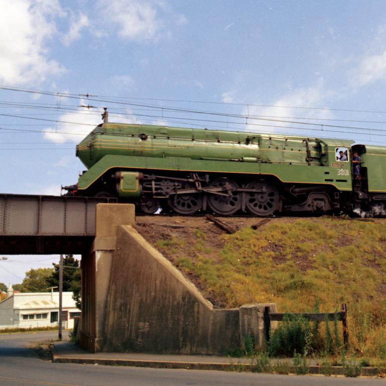A 3801 steam locomotive pulling a passenger train on a raised railway track above a grass embankment. The train is about to cross a railway bridge
