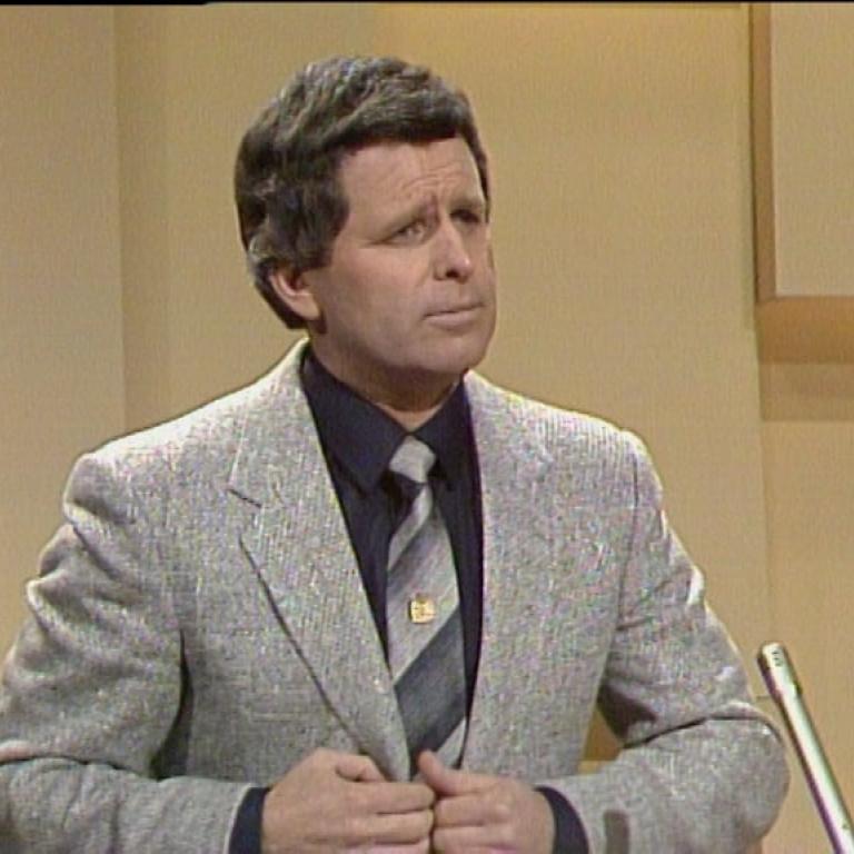 Tony Barber stands in front of a yellow wall wearing a grey suit with a large striped tie.