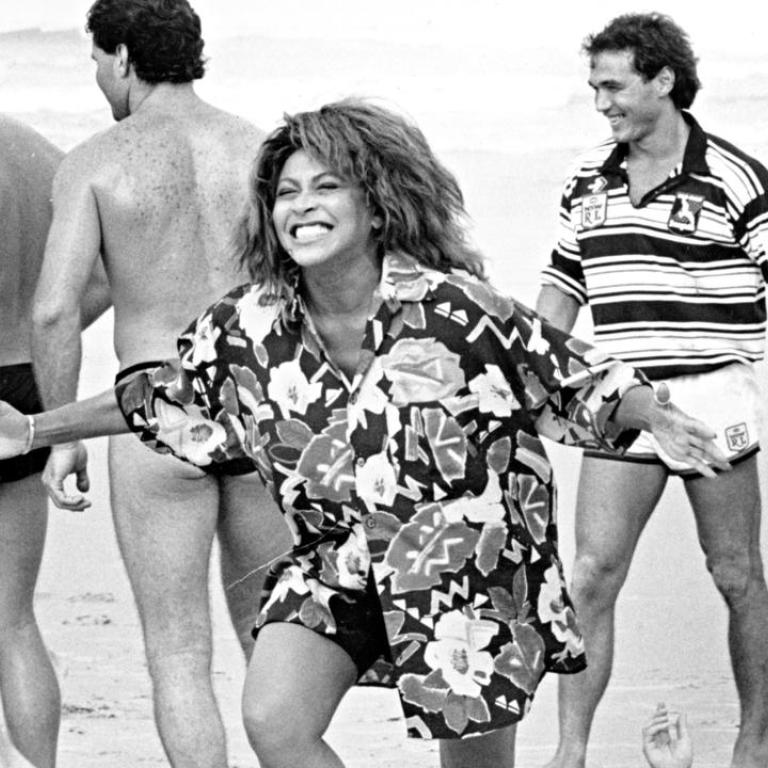 Tina Turner laughing on a beach, surrounded by male rugby league players in swimming costumes.