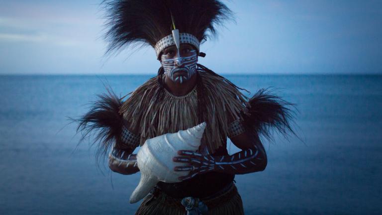 Image featuring Indigenous Francis Williams of the Naygayiw Gigi Dance Troupe in costume standing in front of the sea