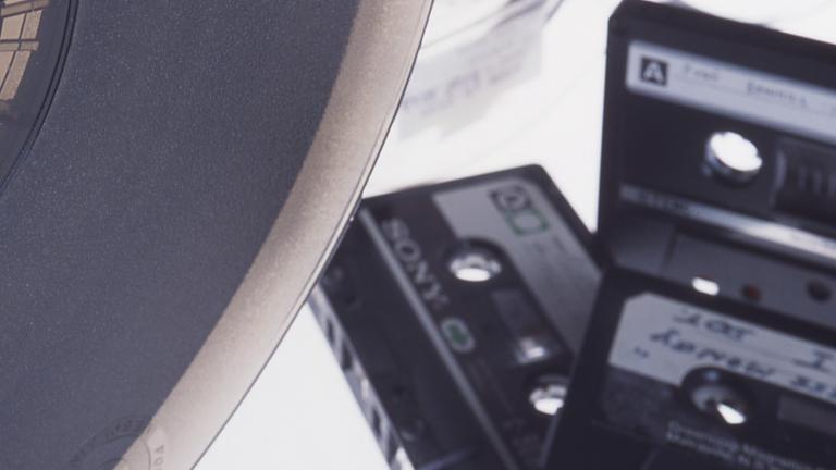 A reel to reel tape is in the foreground in front of three compact cassette tapes