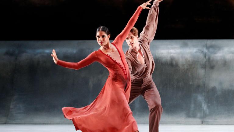 A woman dressed in red and a man dressed in brown dance on a stage against a grey and black backdrop