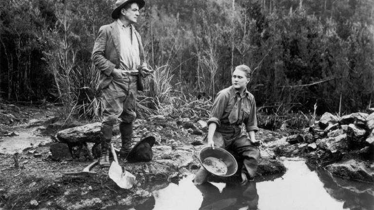Louise Lovely sits by a stream with a gold pan in a scene from Jewelled Nights.