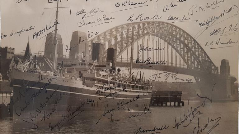 Photo of a ship in front of the Sydney Harbour Bridge. The photo has a lot of signatures on it. 