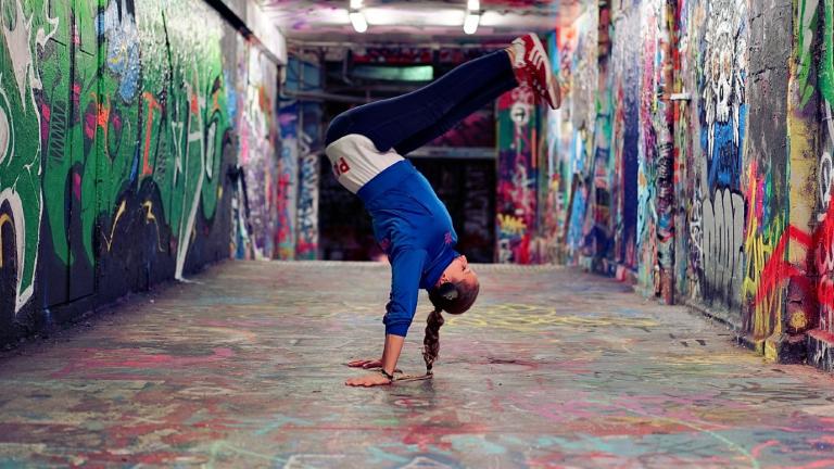 A woman standing on her hands, hip-hop dancing, in a graffitied tunnel