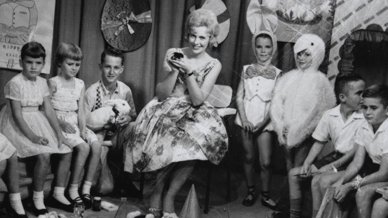 A smiling woman seated in a TV studio surrounded by children. 