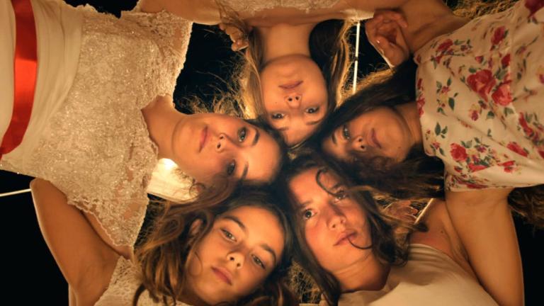 5 young women leaning over linking arms in a circle and looking directly down into the camera.