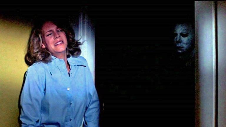 A terrified woman standing against a wall while a shadowy figure in a white mask stands behind her.