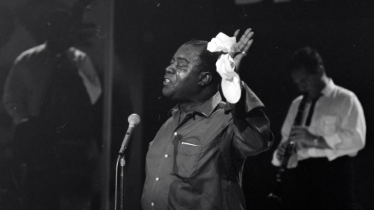 Jazz musician Louis Armstrong standing at a microphone in a TV studio. He has one hand raised and holding a handkerchief. Circa 1964.