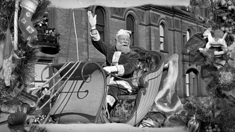An older man dressed as Santa Claus sitting in a sleigh waving at people.