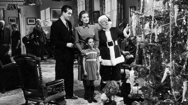 A man, woman and little girl stand with an older man dressed as Santa Claus. They are admiring a Christmas Tree in a large living room with other people gathered in the background.