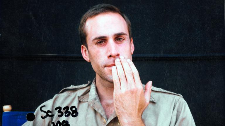 A man pictured from the chest up wearing an army uniform and holding his hand up to his face. His shirt, face and hands are dirty.