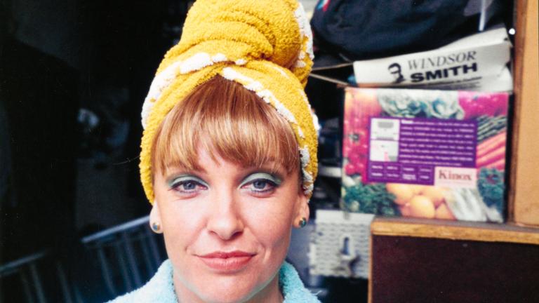 Actor Toni Collette wearing a blue top with her hair wrapped in a yellow towel. Her face is heavily made up with pink lipstick and blue eyeshadow.