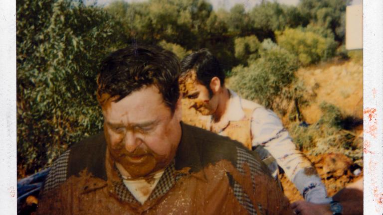 Two men pictured from the chest up walking through some scrubland. They have mud and dirt all over them.
